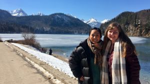 A student, Molly, by a lake with her friend with mountains in the background