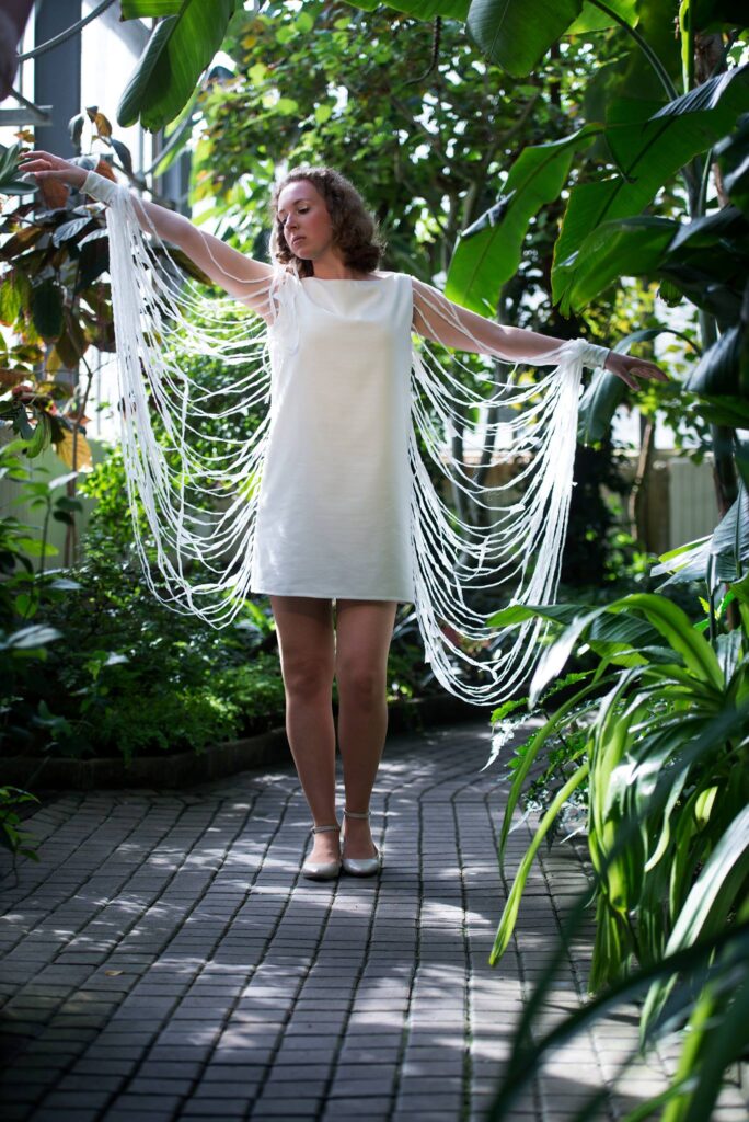 Fashion photography student modelling her own costume project, white dress with botanical garden as background