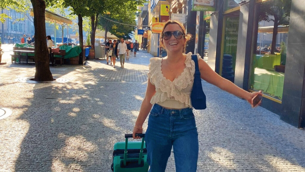 Student smiling with a small roll-on suitcase in the sunny street of Prague.
