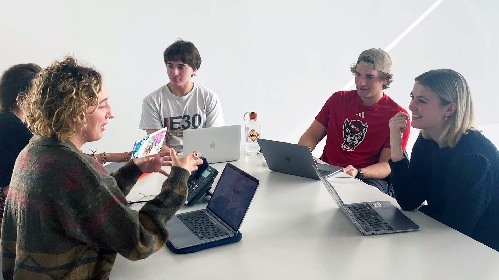 5 students around a table with their laptop computers discussing.