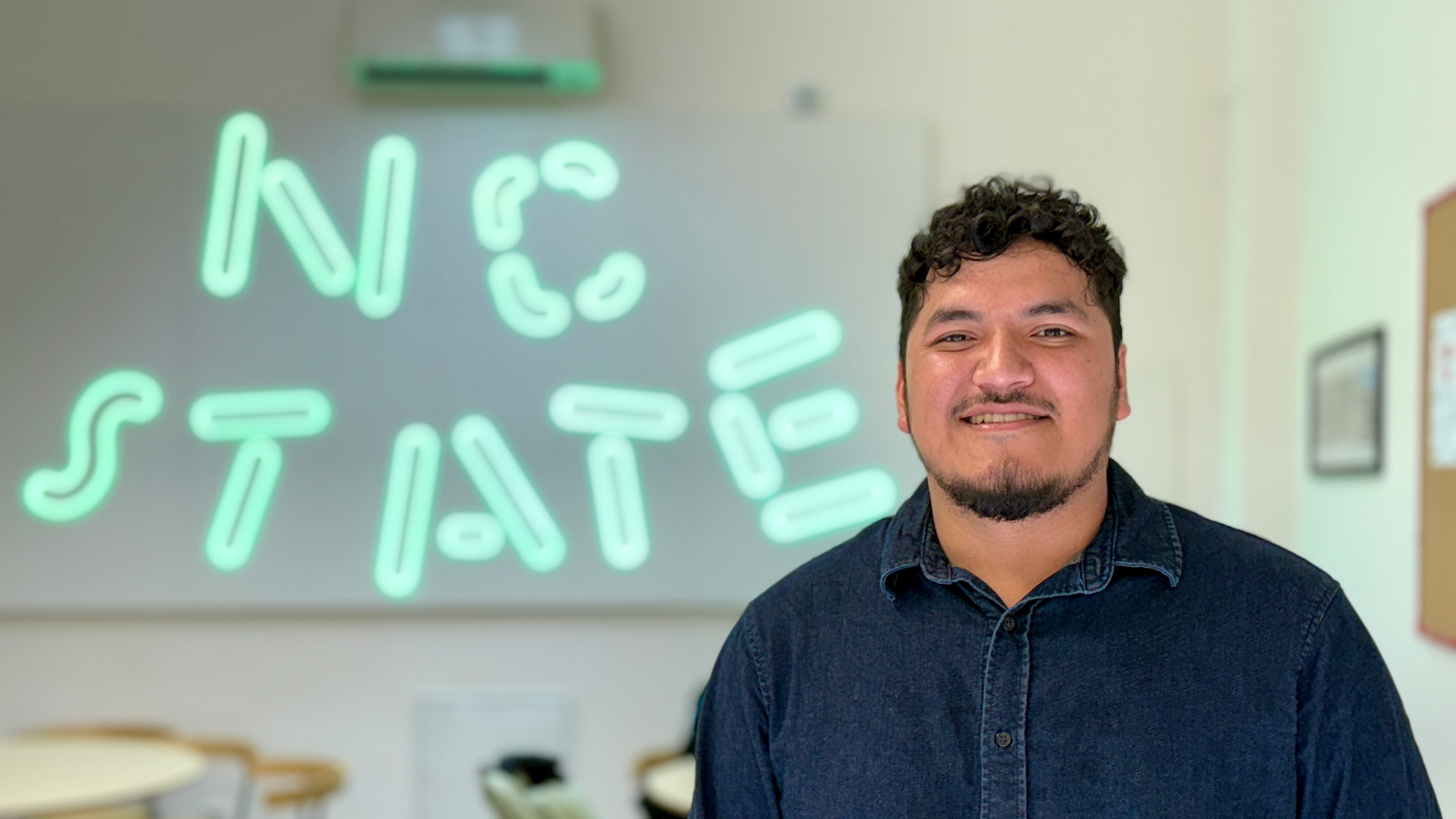 Graduate student in front of a NC State green neon sign.