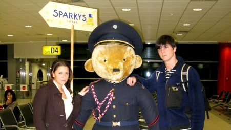 Two students posing for a picture at an airport standing next to a human size plush bear in a uniform